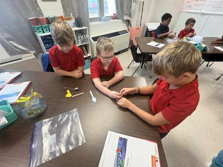 Four children wearing red shirts sit around a table with plastic bags and a book. Behind them are chairs and tables.
