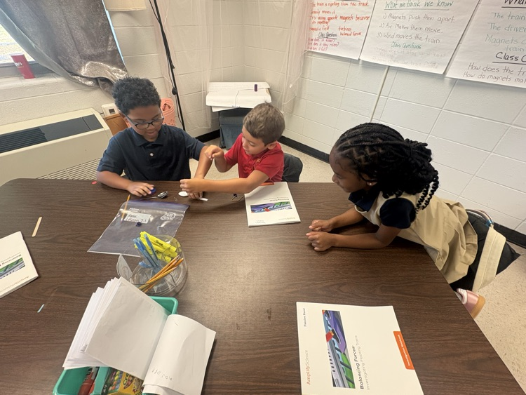 Three children seated at a table in a classroom. They are engaged in a hands-on activity.