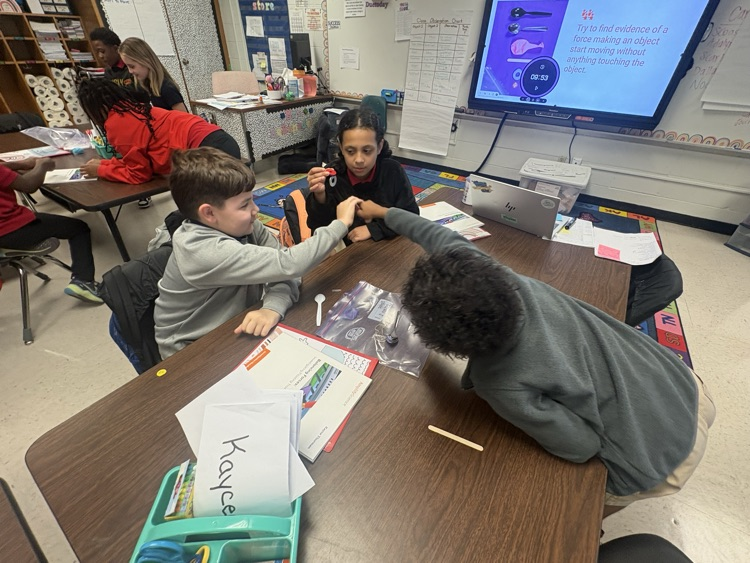 Kids in a classroom, seated around a table, engaged in a learning activity, with a screen in the background displaying information.