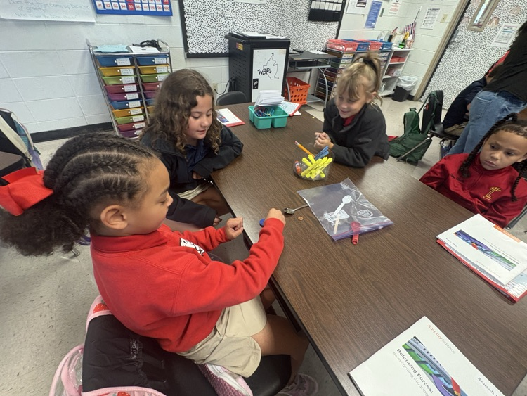 Four children in a classroom setting around a table, engaged in an activity. One child is wearing a red long-sleeve shirt.