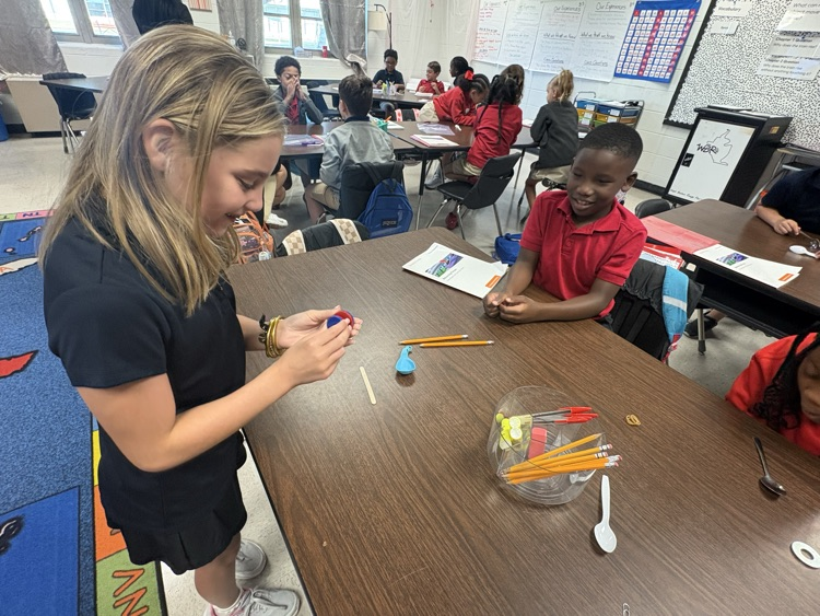 A young girl and boy work together at a table in a classroom, holding small items.