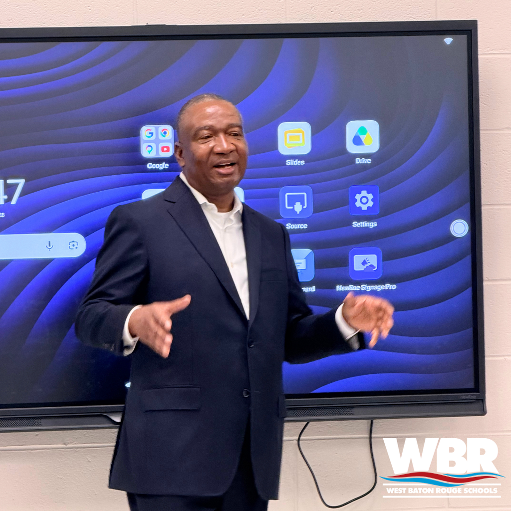Man in a suit stands before a screen displaying a blue wave pattern, talking. Behind him, a white wall.
