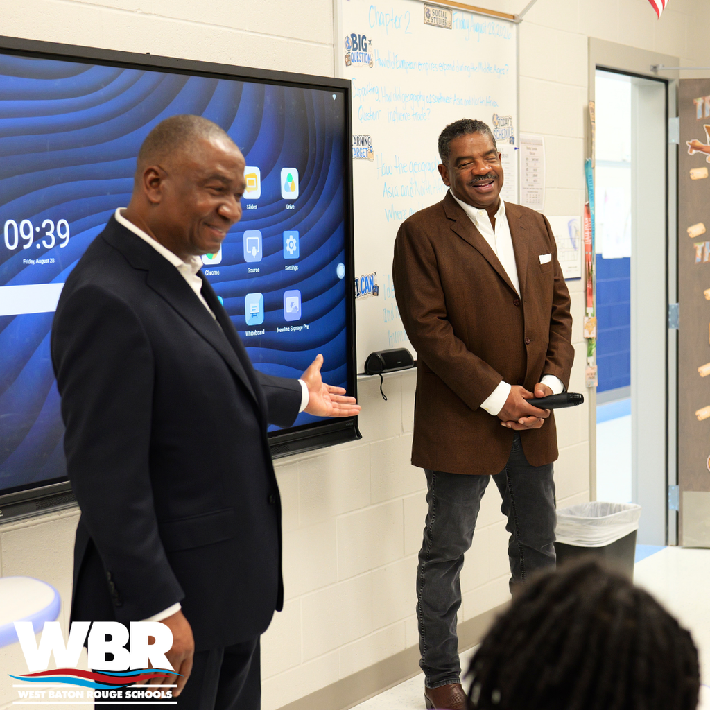 Two men in suits standing in front of a screen with icons and text, one gesturing while smiling.