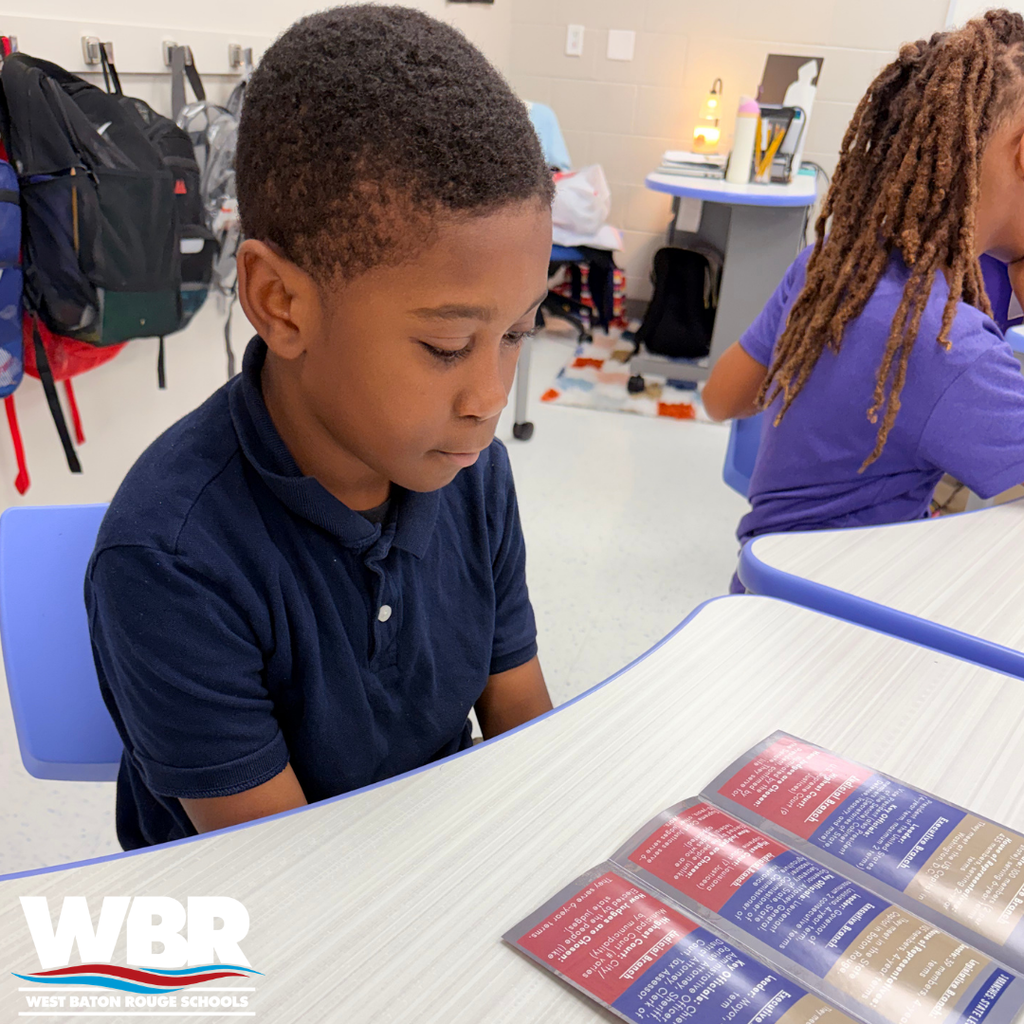 A student wearing a blue polo sits at a desk with a book open, focusing on reading.