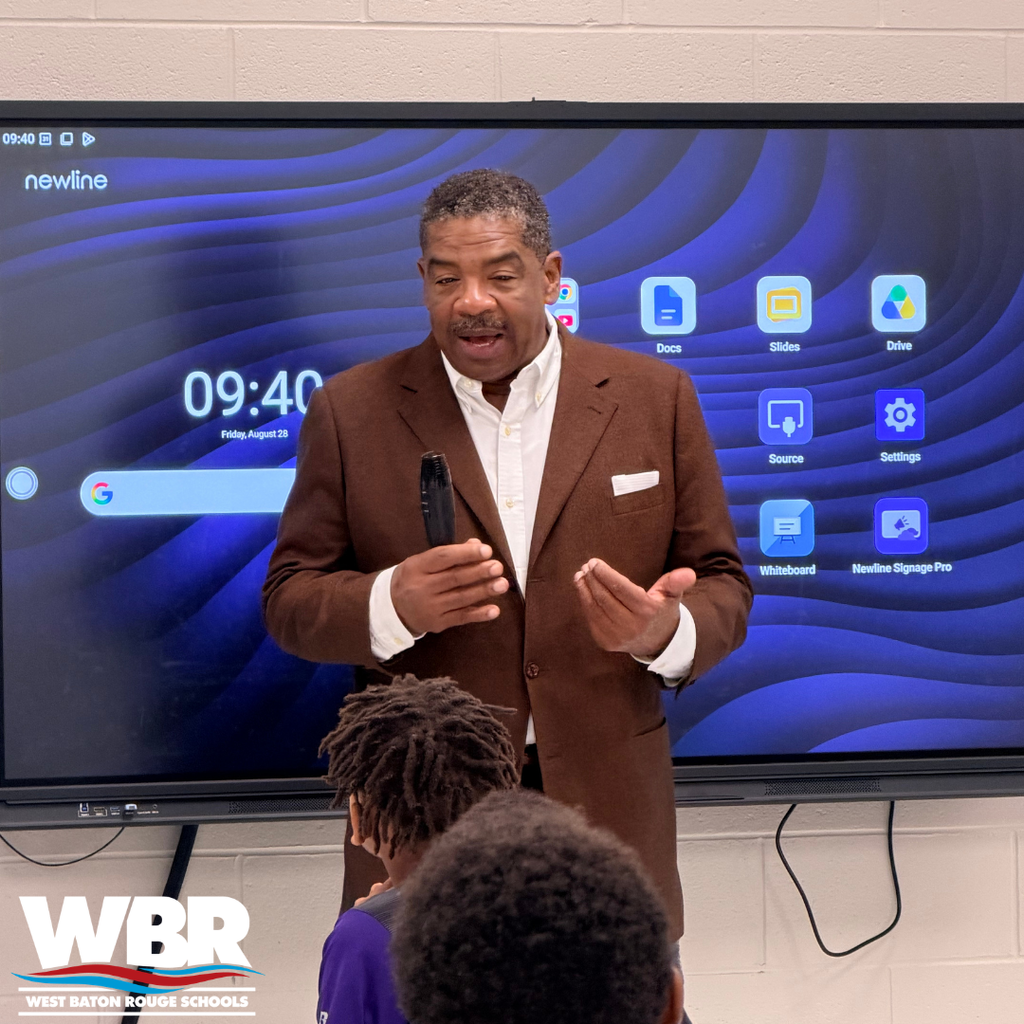 A man in a brown suit speaks to children in front of a screen with blue waves.
