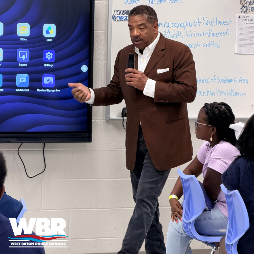 A man in a brown suit speaks into a microphone in a classroom with a digital screen in the background.