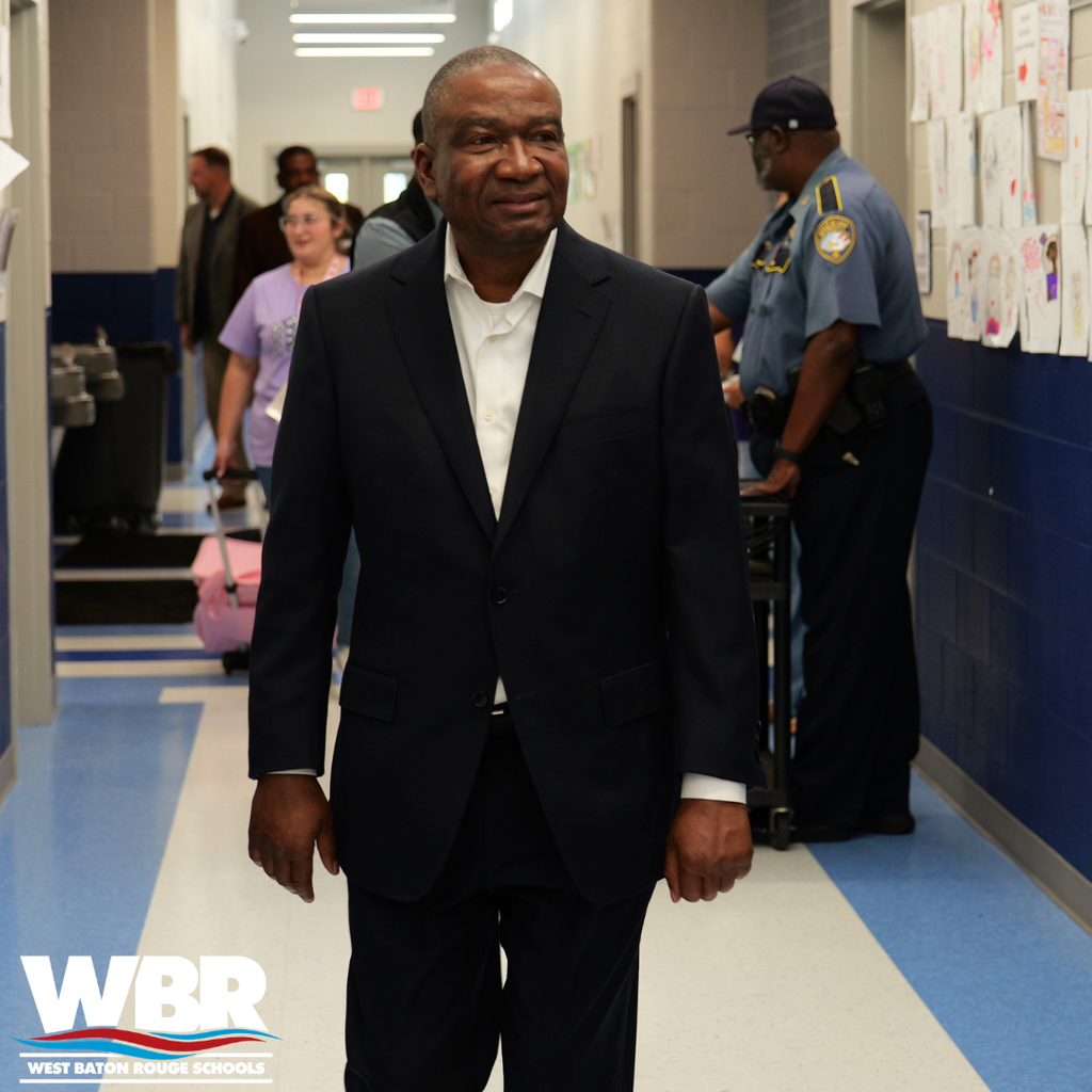 A man in a suit walks down a school hallway with a police officer and others behind.