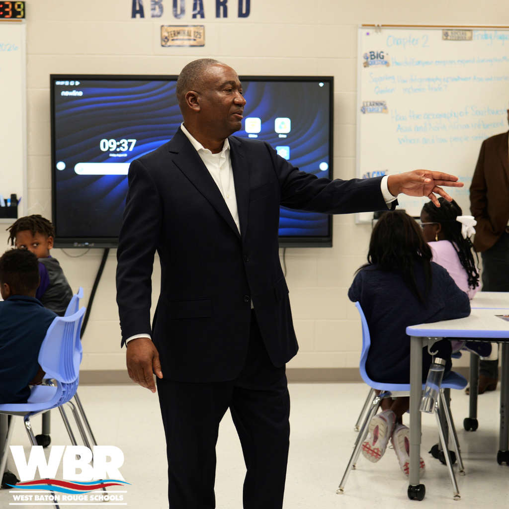 A man in a suit is standing in front of a classroom, gesturing while addressing students.