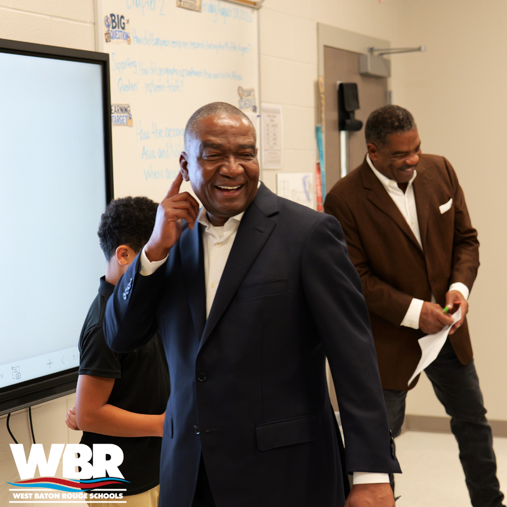 Two men in suits and a child inside a classroom with a large screen on the wall.