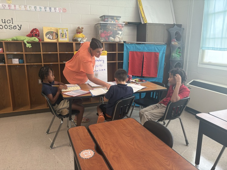 A woman in orange helps three children seated at a desk in a classroom with wooden furniture.