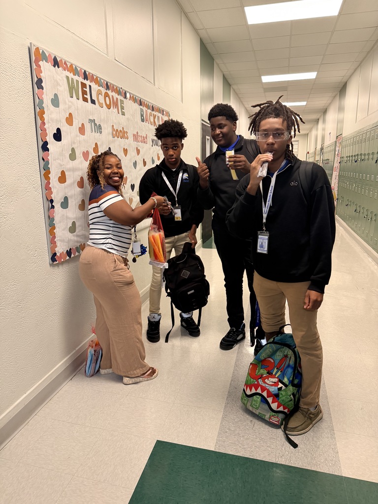 Five people stand in a hallway. Three wear backpacks. A woman shakes hands with a man.