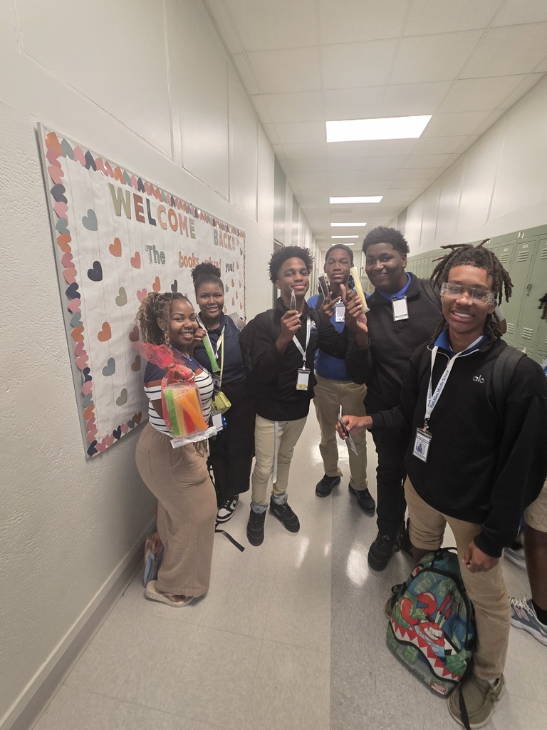 A group of students in a hallway, some holding snacks, standing near a bulletin board with hearts and text.