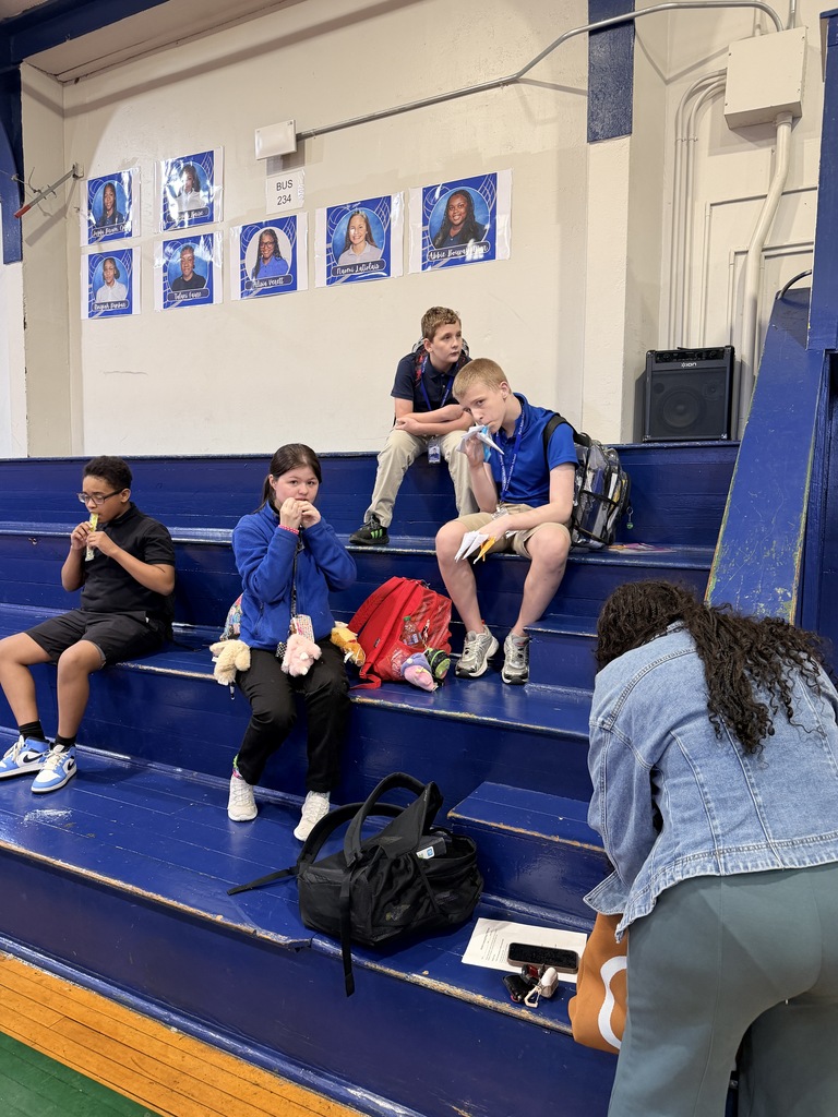 People sitting on blue bleachers in a school gym. One person is bending down, holding a paper.