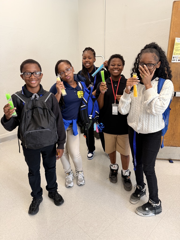 Five children stand together in a corridor, smiling and holding up yellow objects. Two wear glasses, one has a backpack. White walls and a door in the background.