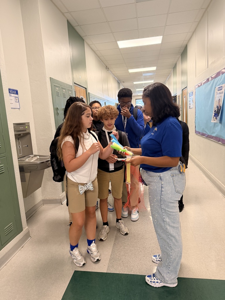 A group of students in a hallway. One student is holding a ribbon. The hallway has white walls, green lockers, and a bulletin board.