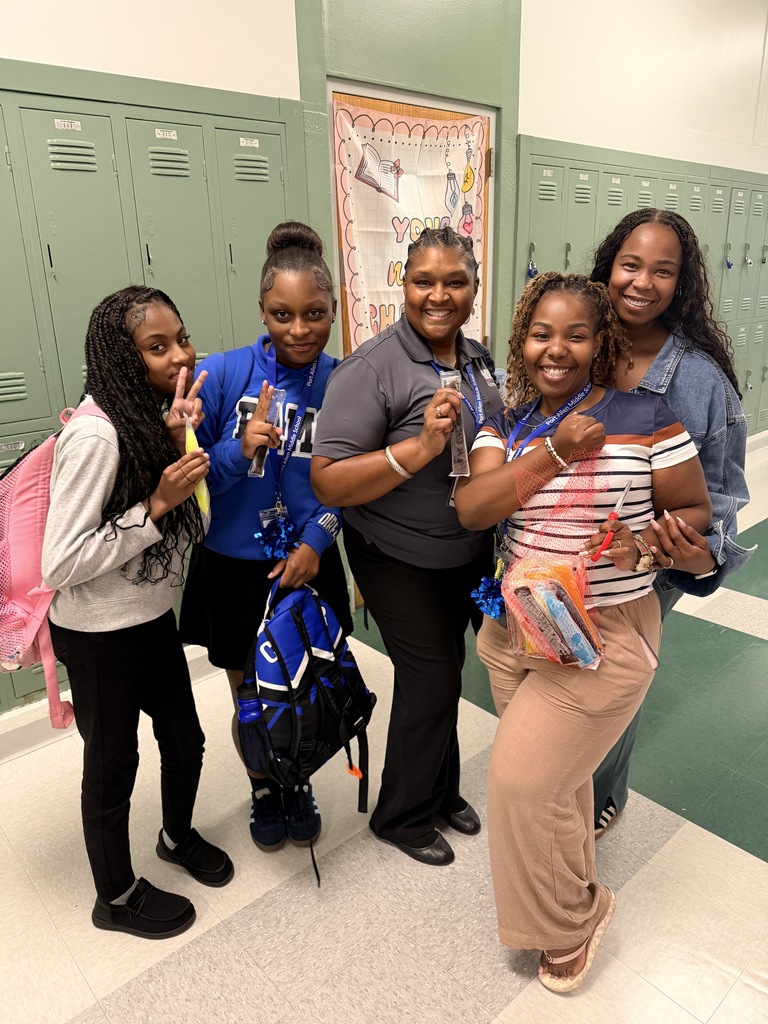 Five women stand close together in a school hallway with lockers in the background. They hold snacks.