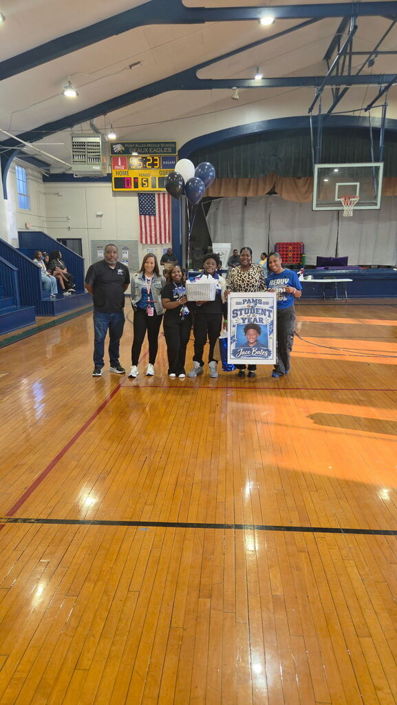 Group of people standing in a gym holding a framed photo and standing next to a banner.