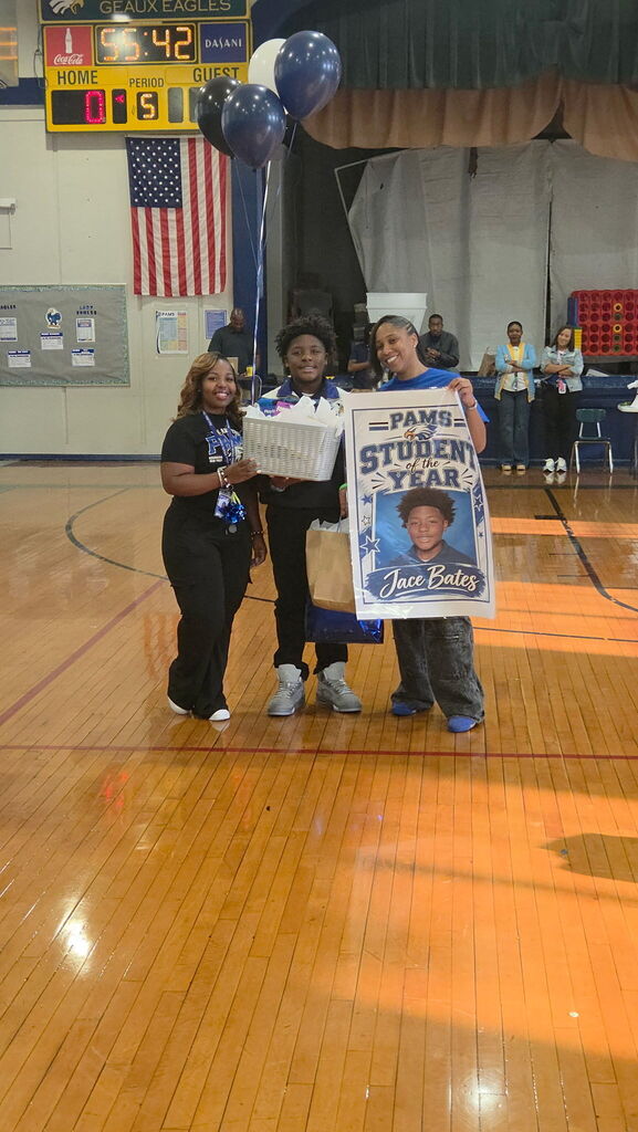 Three individuals stand on a basketball court. A person holds a poster with a photo of a student.