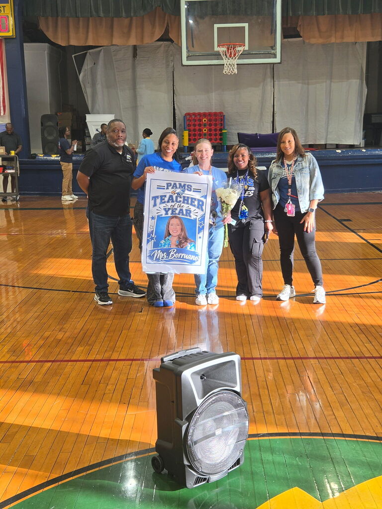 Five adults on a basketball court. A man holds a large sign and bouquet. A speaker sits on the floor.