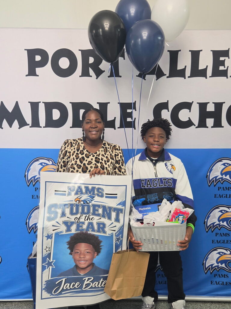 A woman and a child stand by a banner. They hold a sign with "Student of the Year" and gifts.