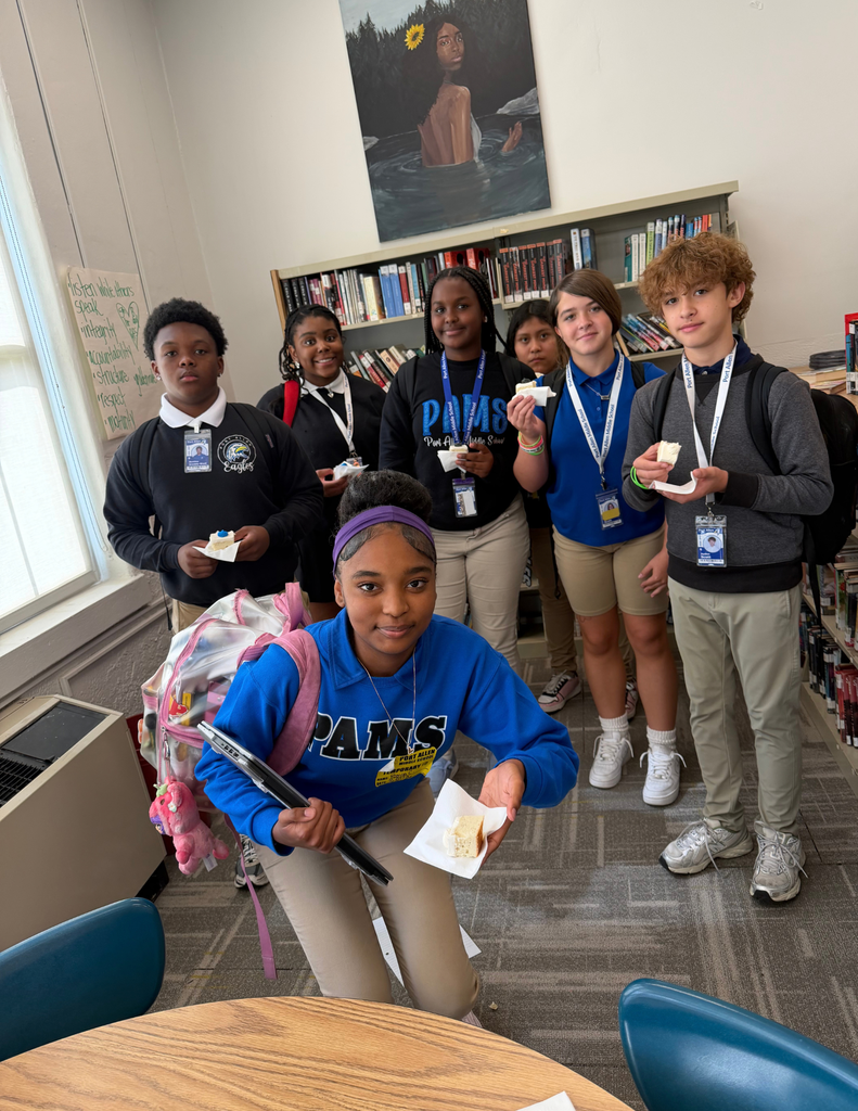 A group of students stand in a library, smiling. They hold small plates of food. A poster hangs on the wall.