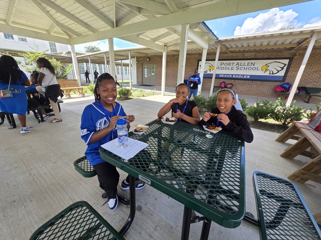 Three children eat at a table under a covered area. A building with a sign is in the background.