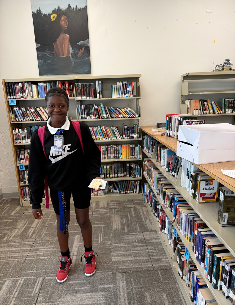 A student in a library wearing a black sweatshirt and red shoes. They hold a small item and stand in front of bookshelves.