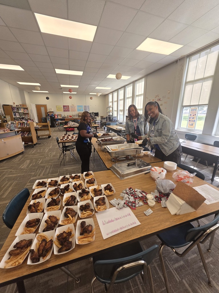 Three people prepare food on a long table in a classroom. Plates with food and bowls are on the table.