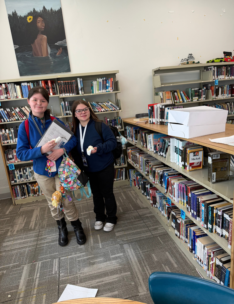 Two individuals stand in a library aisle, one holding a book and a bag, the other with a white lanyard.