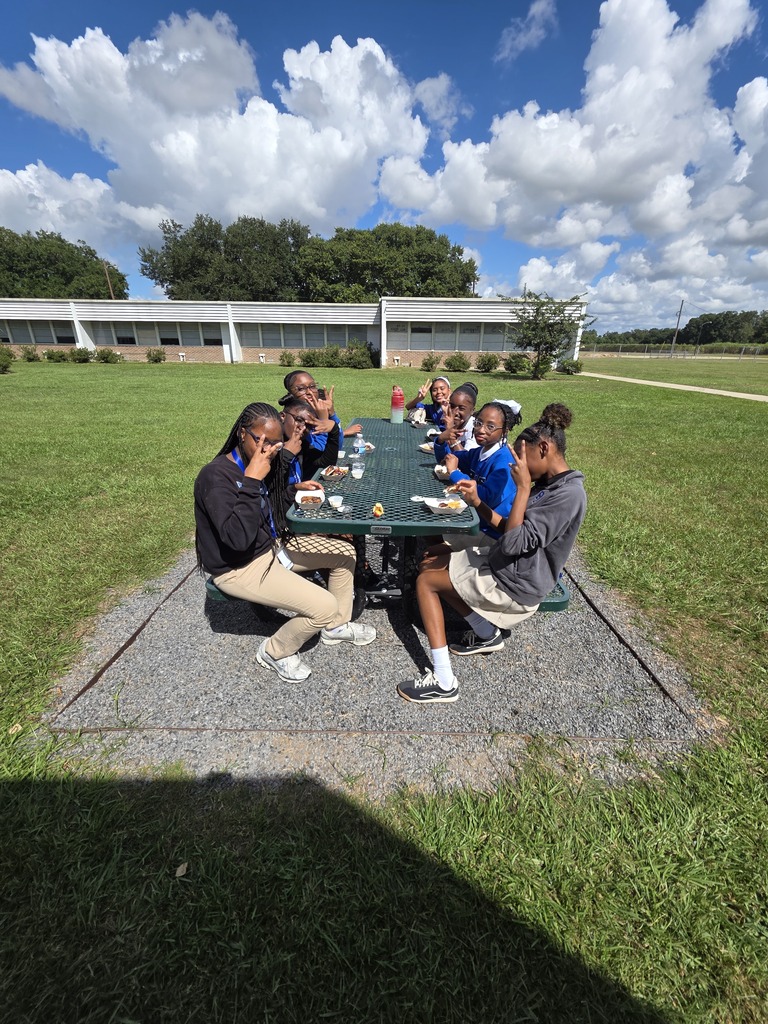 Students sit at a green picnic table in a grassy field, with a school building visible in the background.