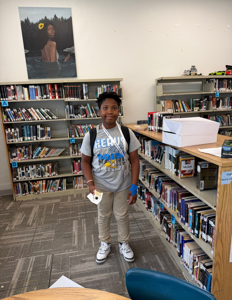 A young person in a library, standing with a book and a backpack. Behind them, shelves filled with books.