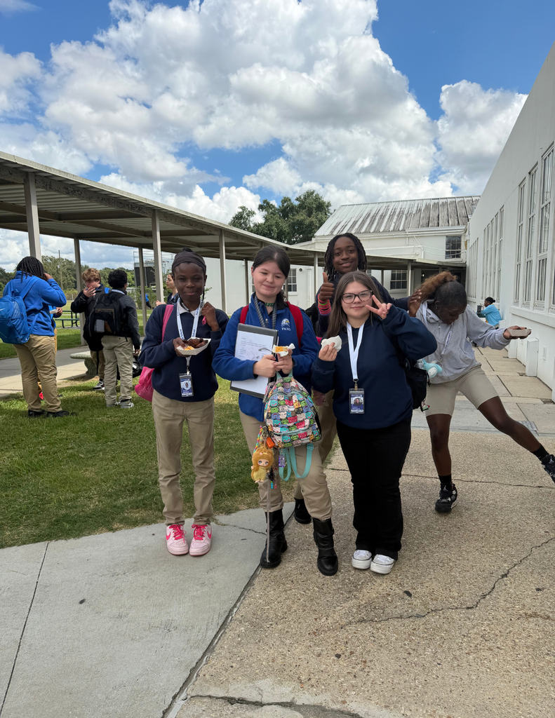 Students pose with food and a certificate outdoors near a school building with a metal roof.