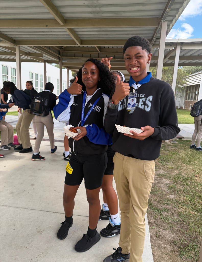 Two students, one in a blue and black jacket, the other in black, smile while holding food. Behind them, others stand on a paved area.