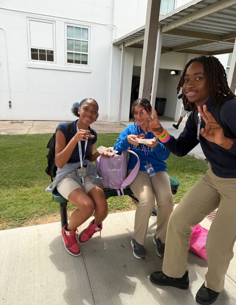 Three students sitting on a bench outdoors with one making peace sign; a white building with windows in background.