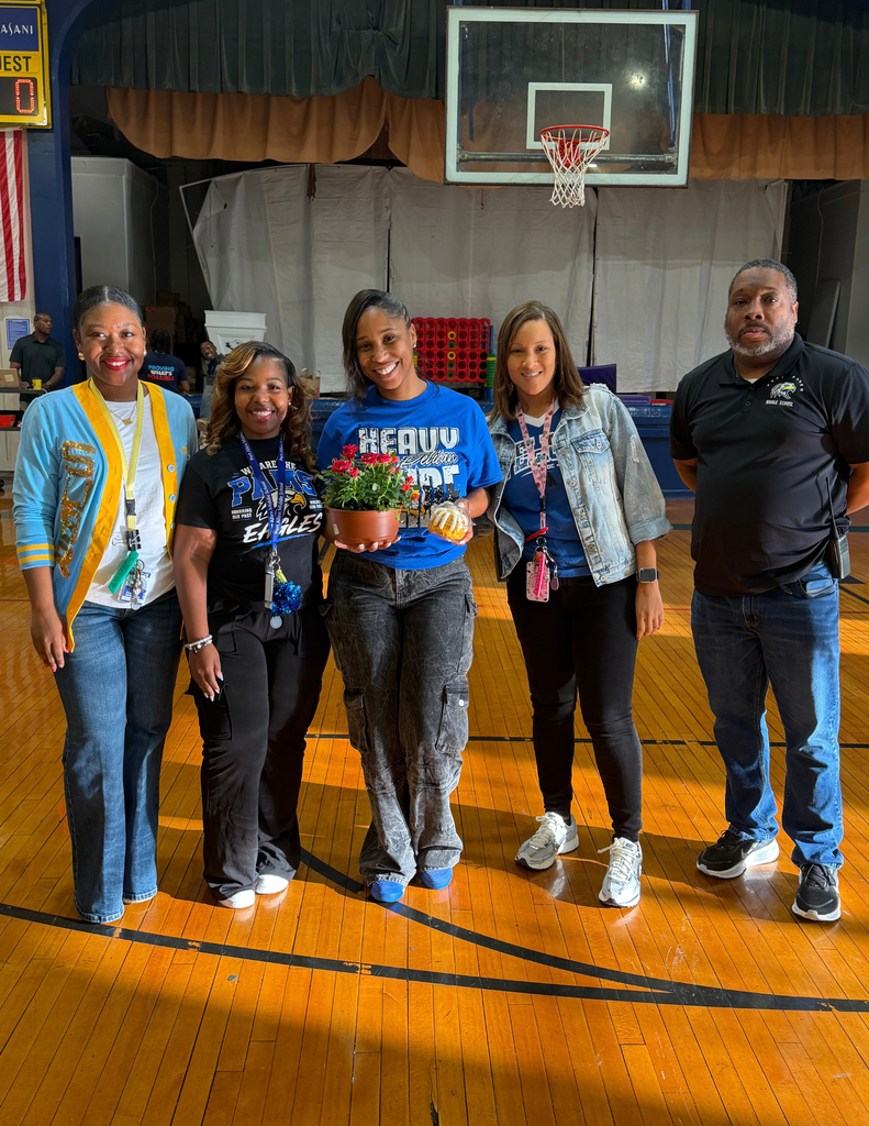Five individuals stand in a basketball court with a basketball hoop in the background. One person holds a potted plant.