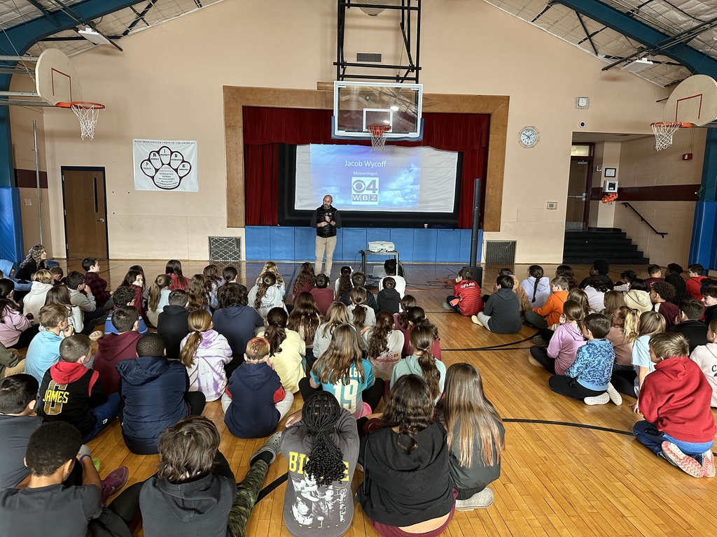 A man in a puffy black vest speaks to students seated on a gym floor.