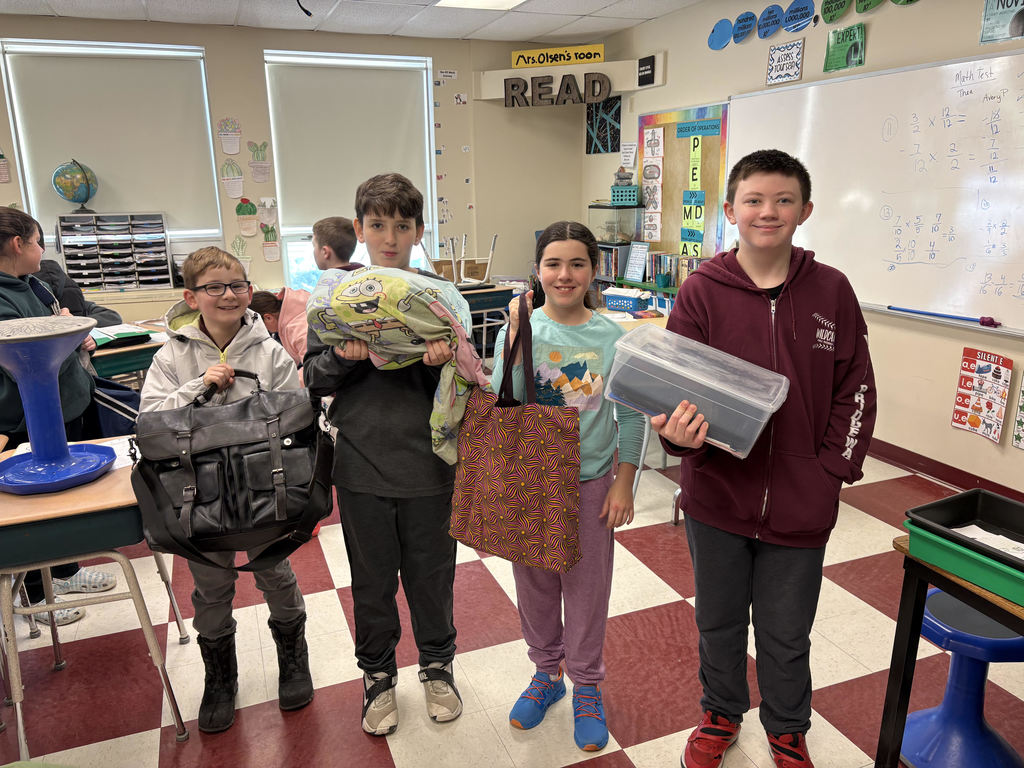Four students hold up containers they used instead of their backpacks for a fun "anything but a backpack:" spirit day. There are briefcases, pillowcase, totebags, and rubbermaid bins.