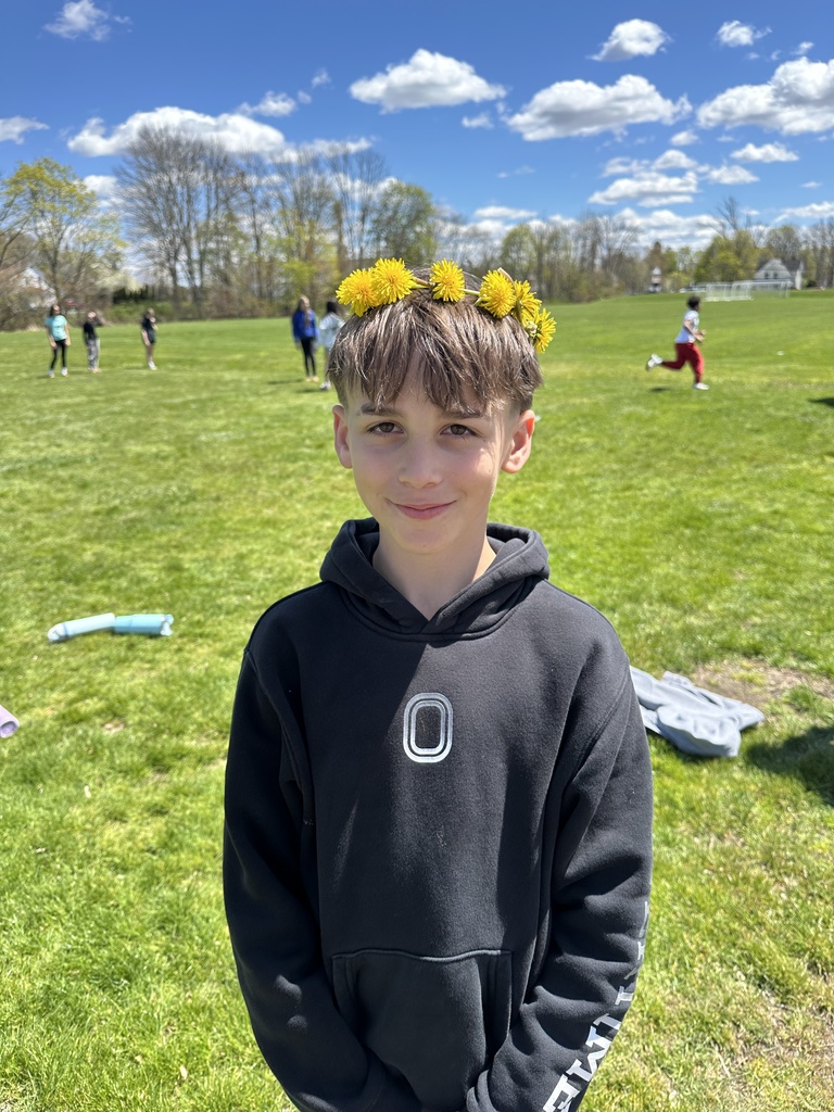 a boy wears a dandelion flower crown in front of a green field and blue sky with fluffy clouds.  Children play in the background.  