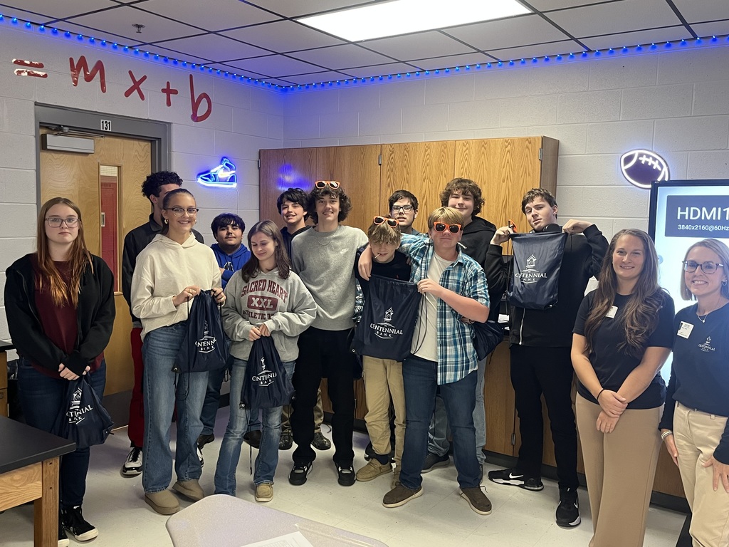 A group of students and Centennial Bank presenters stands in a classroom. Several students are holding drawstring bags with the Centennial Bank logo.