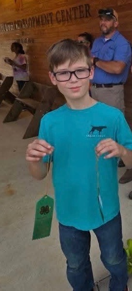 boy in glasses, a turquoise T-shirt, and jeans holds up two green 4-H ribbons and smiles at the camera. Other participants and adults are visible in the background at the event.