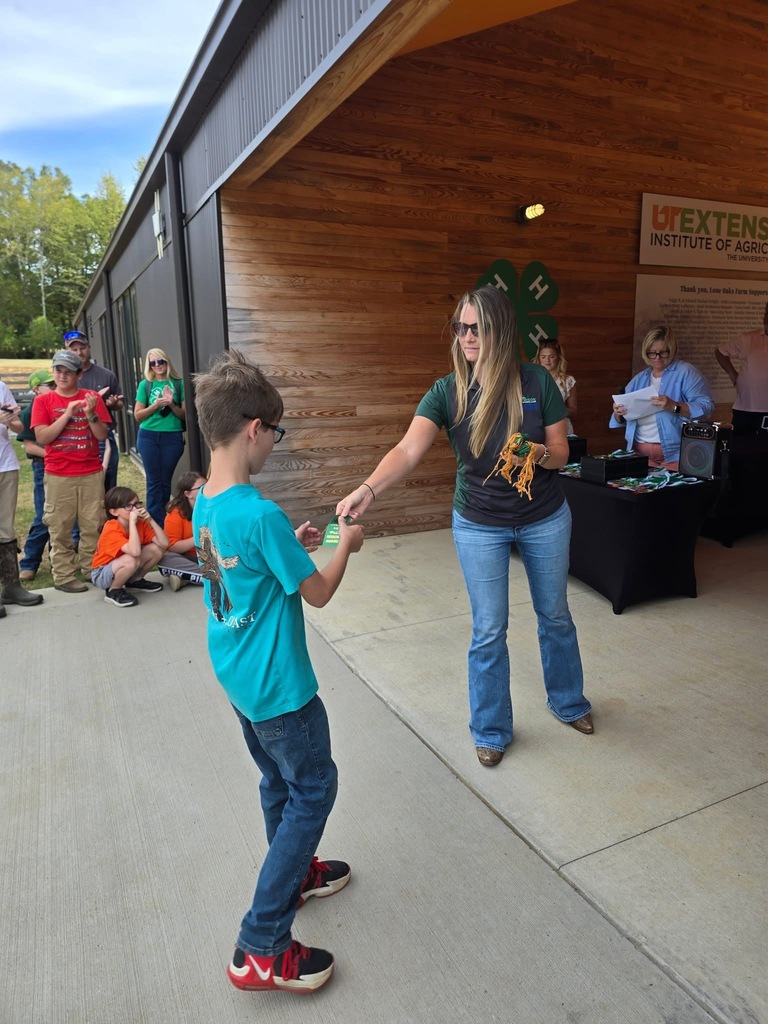 A woman in jeans and sunglasses hands an award ribbon to a boy wearing glasses, a turquoise T-shirt, and jeans at a 4-H event. People stand and clap in the background near a UT Extension Institute of Agriculture booth.