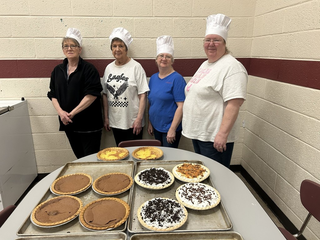 four wces lunch ladies standing behing table full of pies wearing chef hats