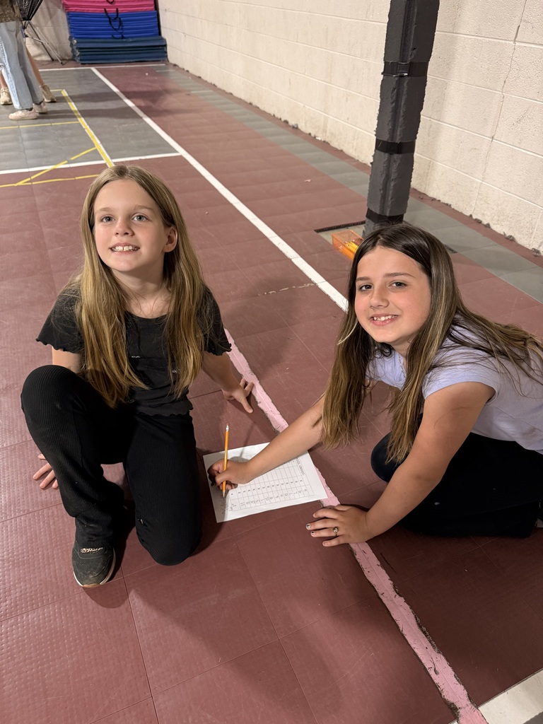 Two students kneel on the gym floor beside a Frisbee Math worksheet while recording answers with a pencil.