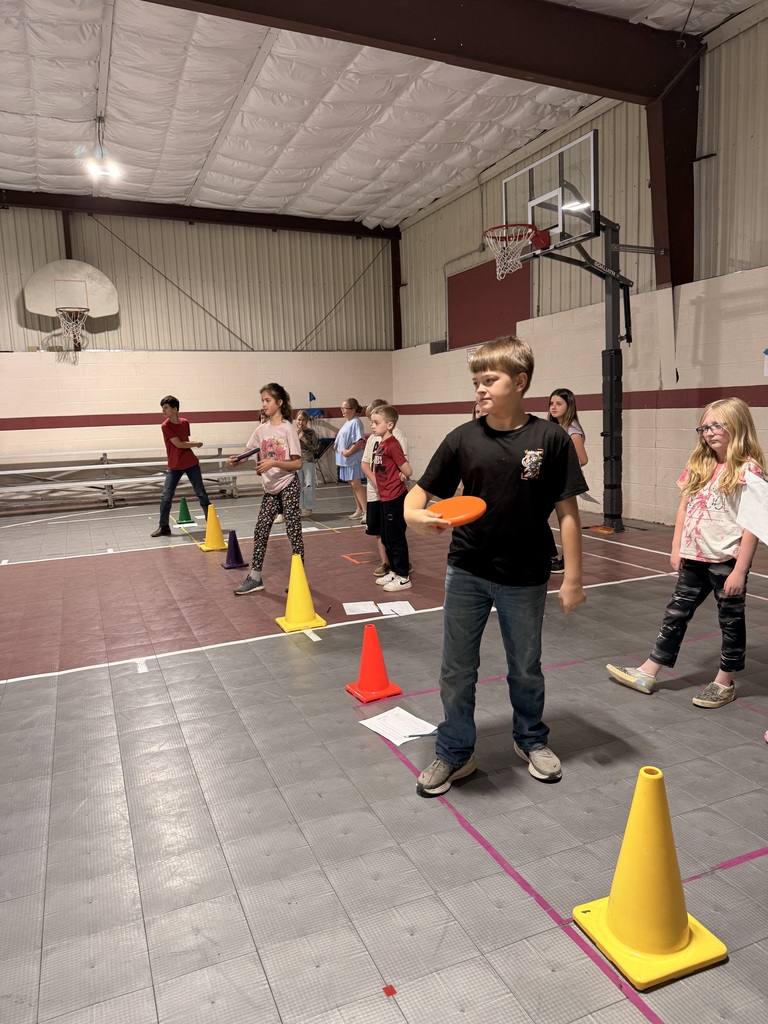 Students stand in lines in the gym during a multiplication frisbee activity, with cones and worksheets placed on the floor.