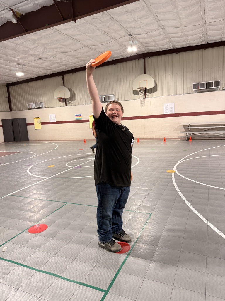 A student smiles while standing in the gym and holding an orange frisbee above his head.
