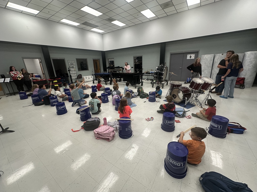 West Carroll Little Drummers Clinic with elementary students in grades K-4 seated in the band room with blue buckets and drumsticks. High school musicians and instructors are positioned around the room with band and percussion instruments as the students learn bucket drumming.
