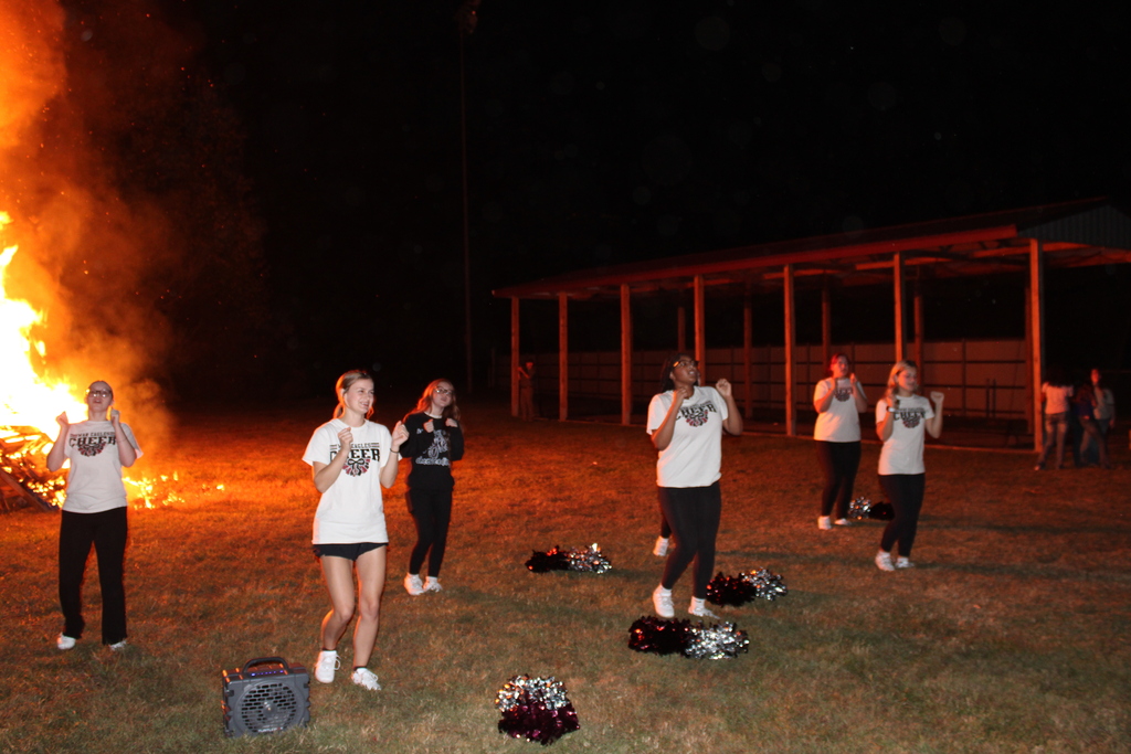 Series of photos from the West Carroll Homecoming Bonfire held at night on school grounds. Students and staff gather with friends around a large bonfire, pose for group photos, and enjoy the Homecoming celebration. West Carroll cheerleaders perform near the fire and serve refreshments, while additional photos show the bonfire being prepared and lit with safety personnel nearby.