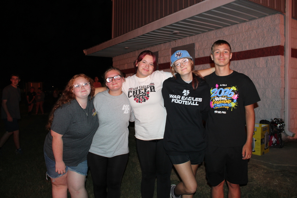 Series of photos from the West Carroll Homecoming Bonfire held at night on school grounds. Students and staff gather with friends around a large bonfire, pose for group photos, and enjoy the Homecoming celebration. West Carroll cheerleaders perform near the fire and serve refreshments, while additional photos show the bonfire being prepared and lit with safety personnel nearby.