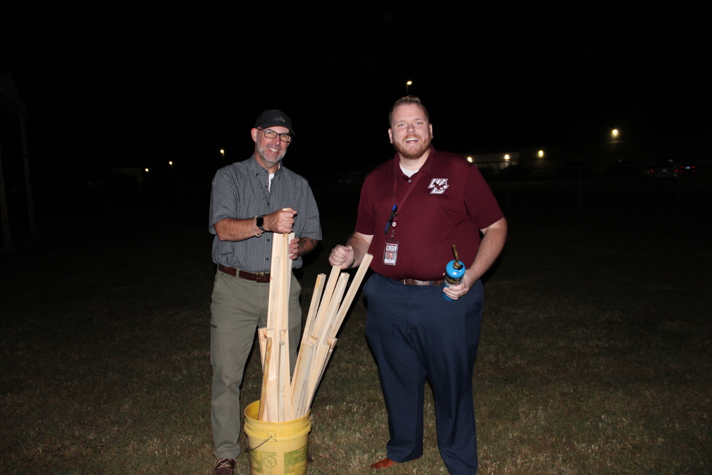Series of photos from the West Carroll Homecoming Bonfire held at night on school grounds. Students and staff gather with friends around a large bonfire, pose for group photos, and enjoy the Homecoming celebration. West Carroll cheerleaders perform near the fire and serve refreshments, while additional photos show the bonfire being prepared and lit with safety personnel nearby.