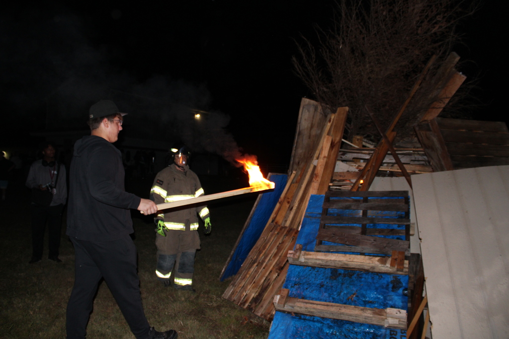Series of photos from the West Carroll Homecoming Bonfire held at night on school grounds. Students and staff gather with friends around a large bonfire, pose for group photos, and enjoy the Homecoming celebration. West Carroll cheerleaders perform near the fire and serve refreshments, while additional photos show the bonfire being prepared and lit with safety personnel nearby.